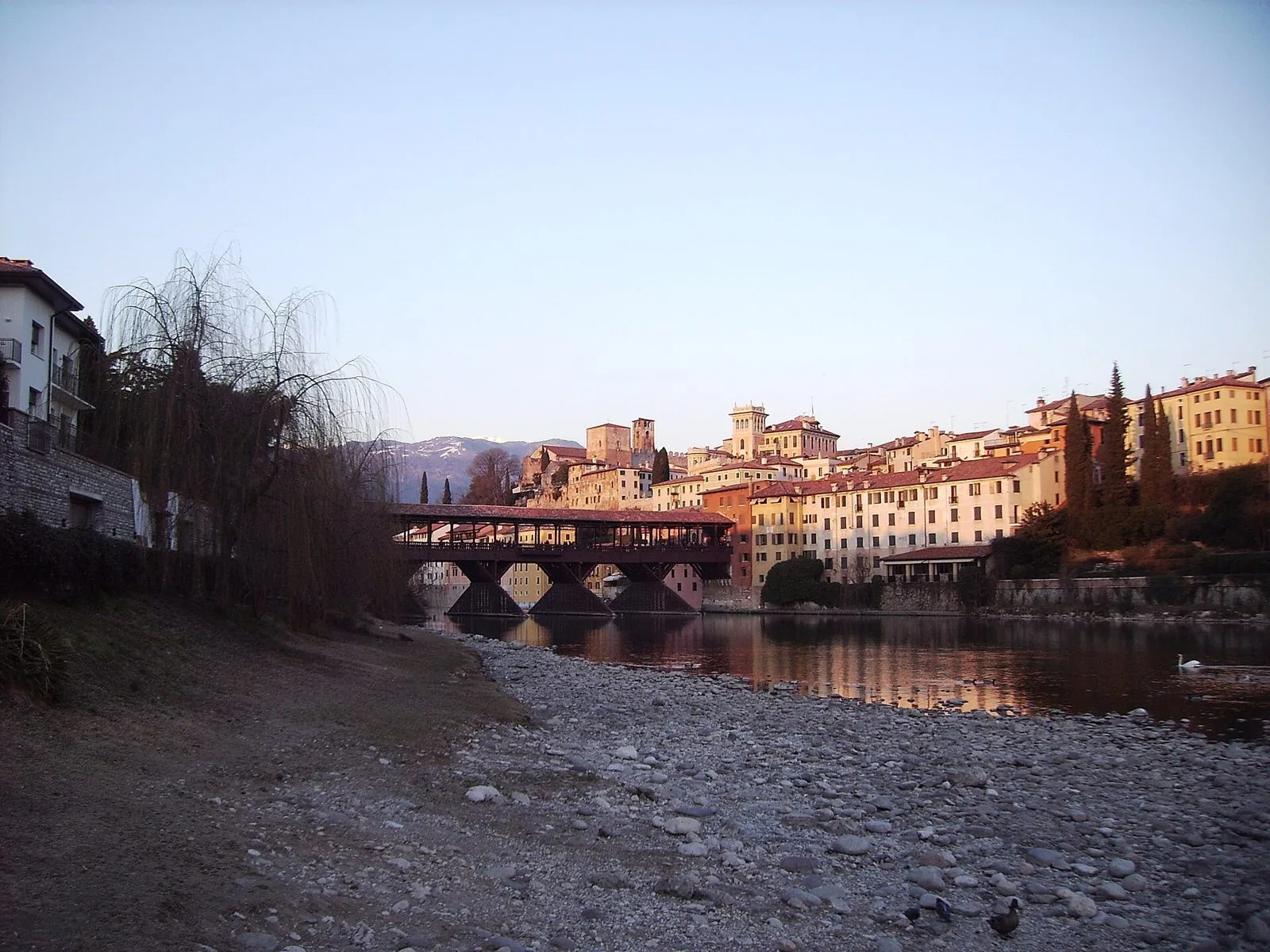 Il Ponte Vecchio di Bassano del Grappa e le case del centro storico illuminate dalla luce calda di una sera d'inverno, con le montagne innevate sullo sfondo