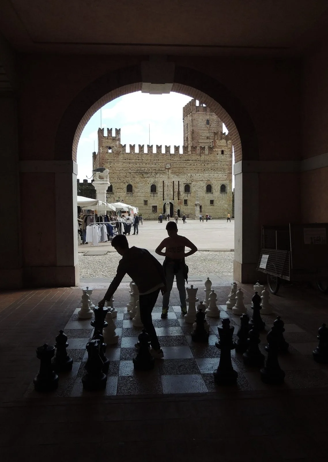 Due ragazzi in controluce giocano con grandi pezzi degli scacchi sotto un portico di Marostica; sullo sfondo, oltre l'arco, la piazza e il Castello Inferiore merlato