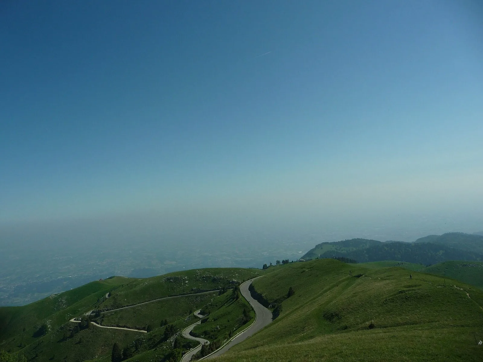 Strada di montagna a tornanti tra i prati verdi del Monte Grappa, con la pianura veneta velata di foschia sullo sfondo